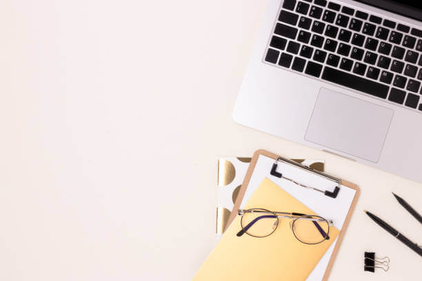 photo d'un bureau vu du dessus où se trouve un ordinateur, un bloc note, un cahier, des lunettes de vue, des stylos et une pince